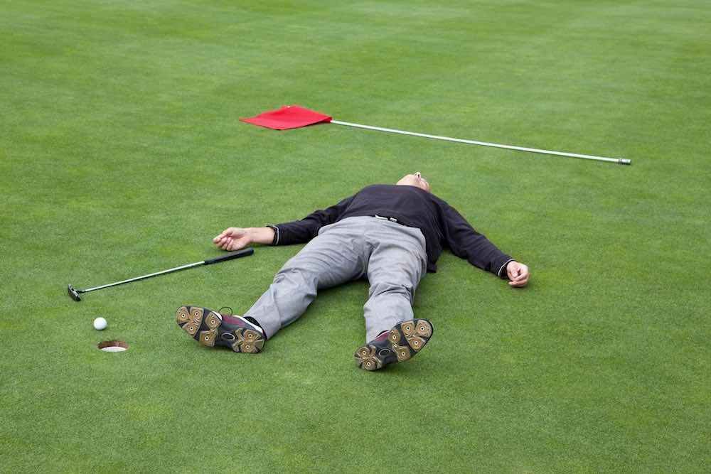 A distraught golfer lying on putting green with ball at the edge of hole