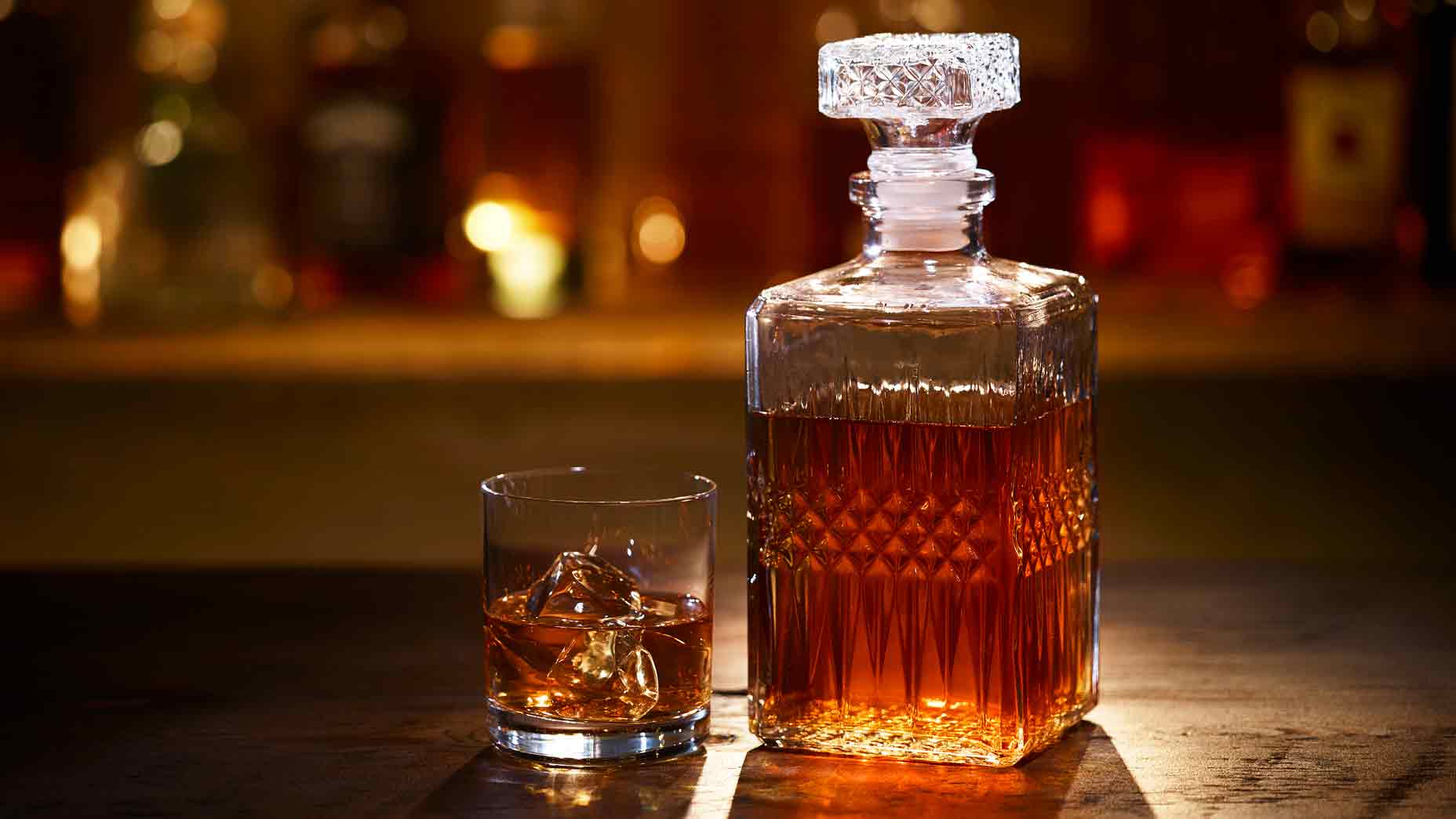 A Bourbon Whiskey Decanter and Bourbon on the Rocks in a Tumbler sitting on a bar with bottles in the background.