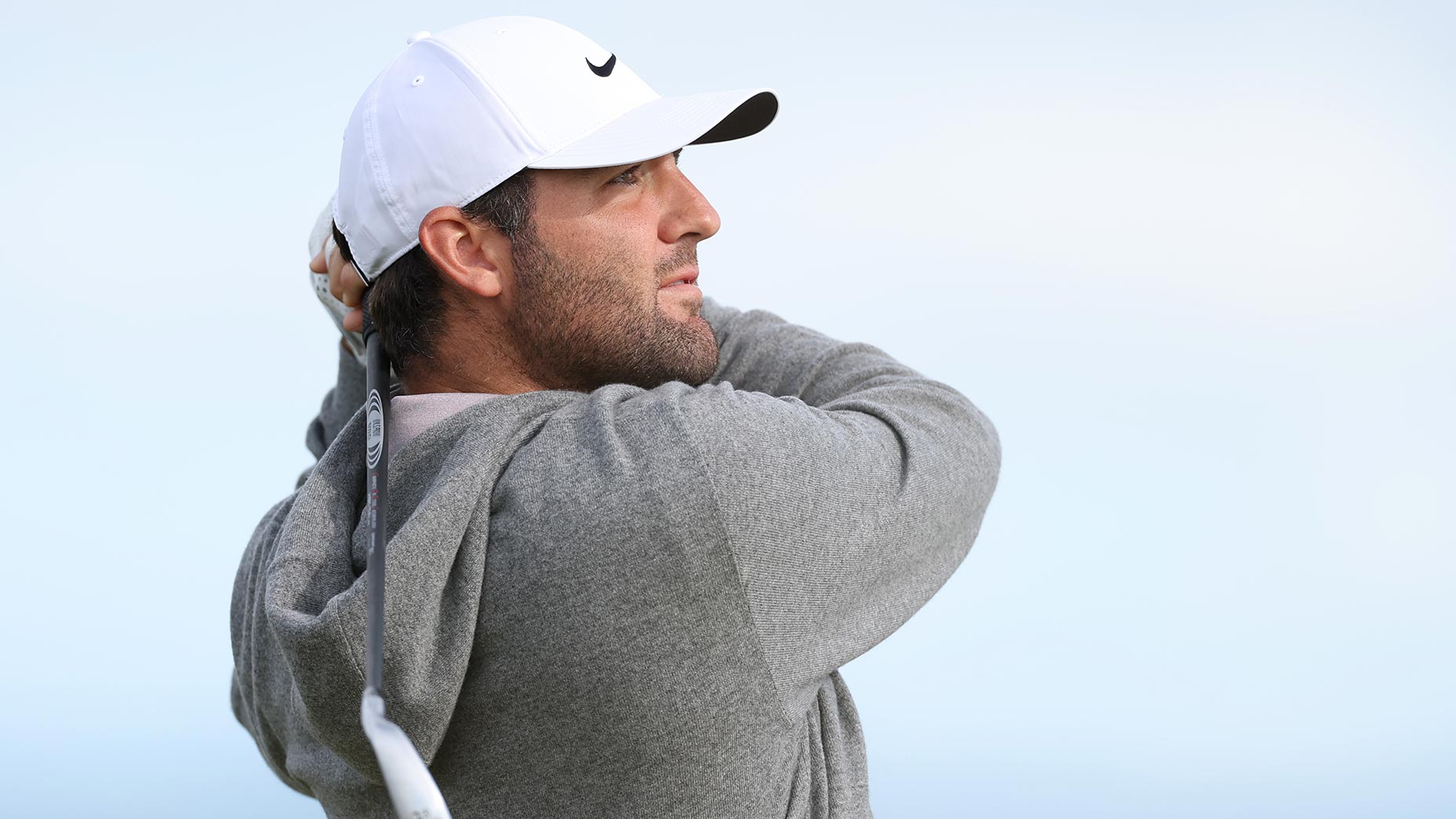 scottie scheffler swings iron at the genesis scottish open in gray sweatshirt and white hat