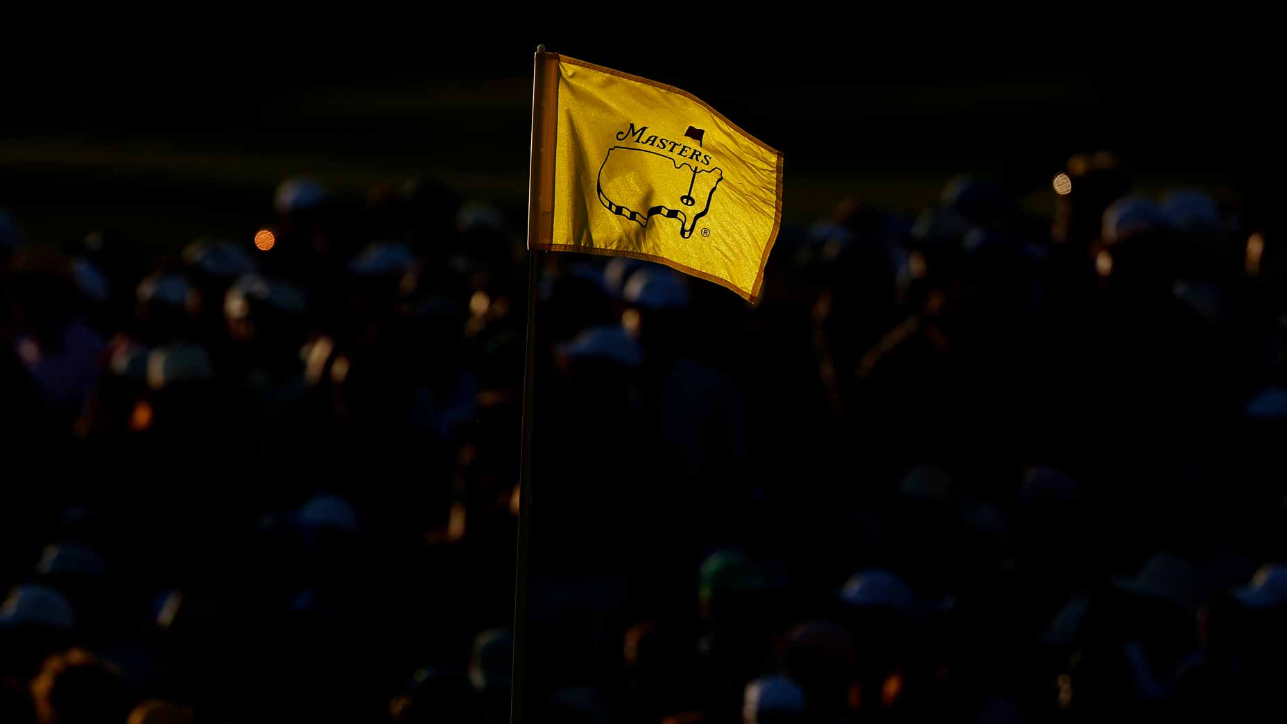 A yellow 2026 Masters golf tournament flag waves in the foreground, illuminated against a dark, blurry background filled with silhouettes of people.