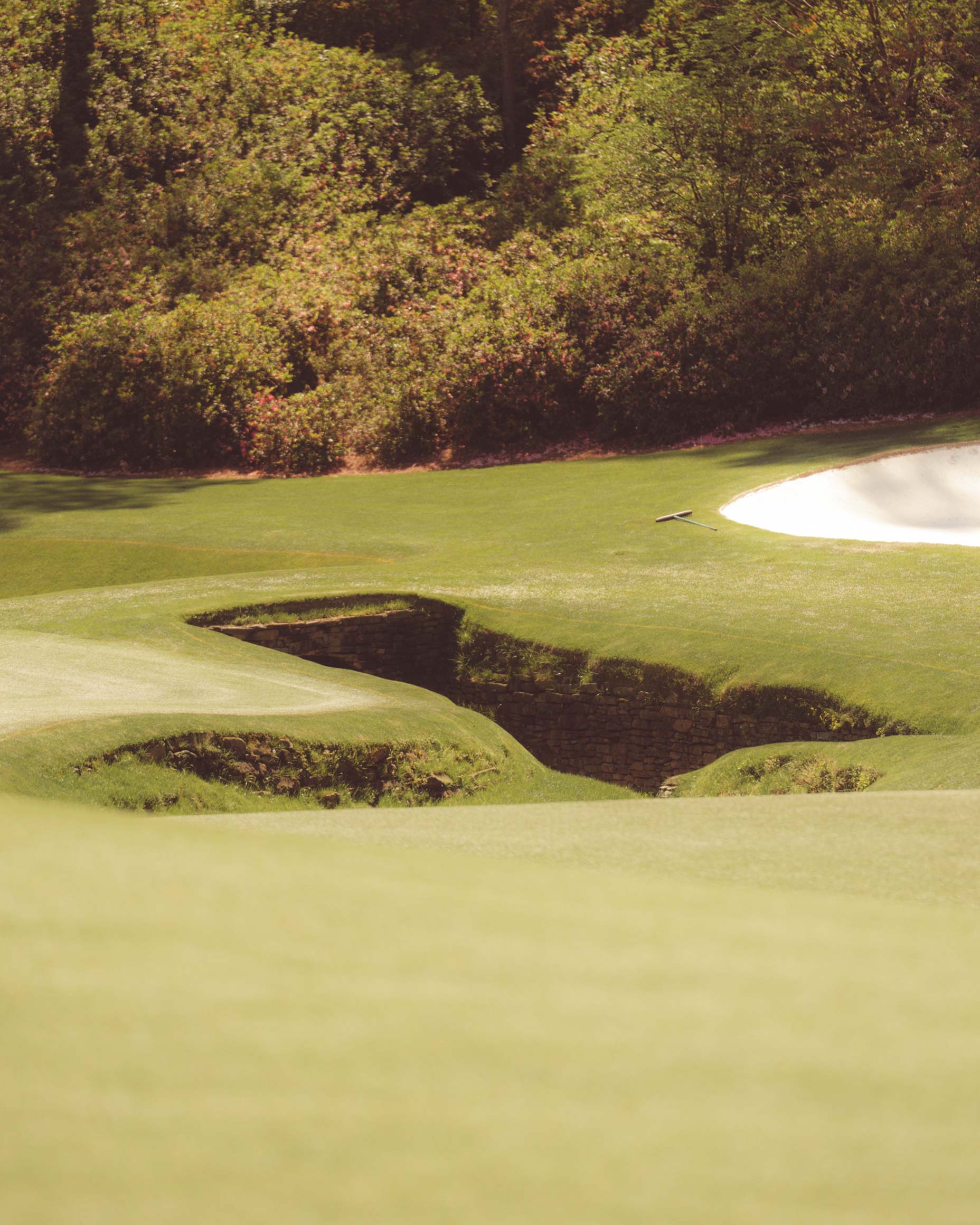 A golf course with a green fairway, a sand bunker on the right, and a small stone-lined creek running through the grass—reminiscent of Augusta National—surrounded by dense trees and shrubbery in the background.
