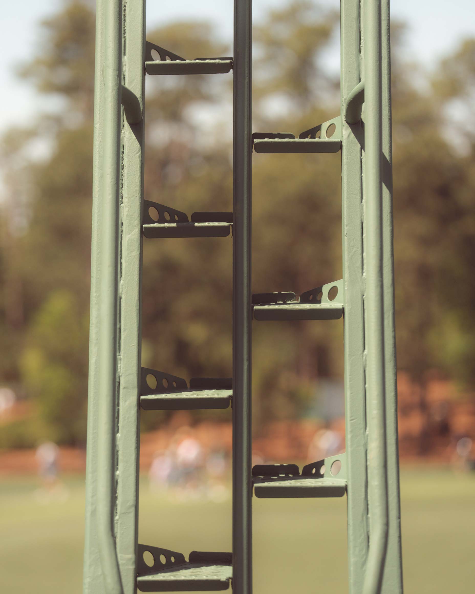Close-up of a green metal ladder-like structure, reminiscent of Augusta National, with trees and a blurry sports field in the background. The focus is on the metal bars, echoing the feeling after you've walked all 18 at a classic golf course.