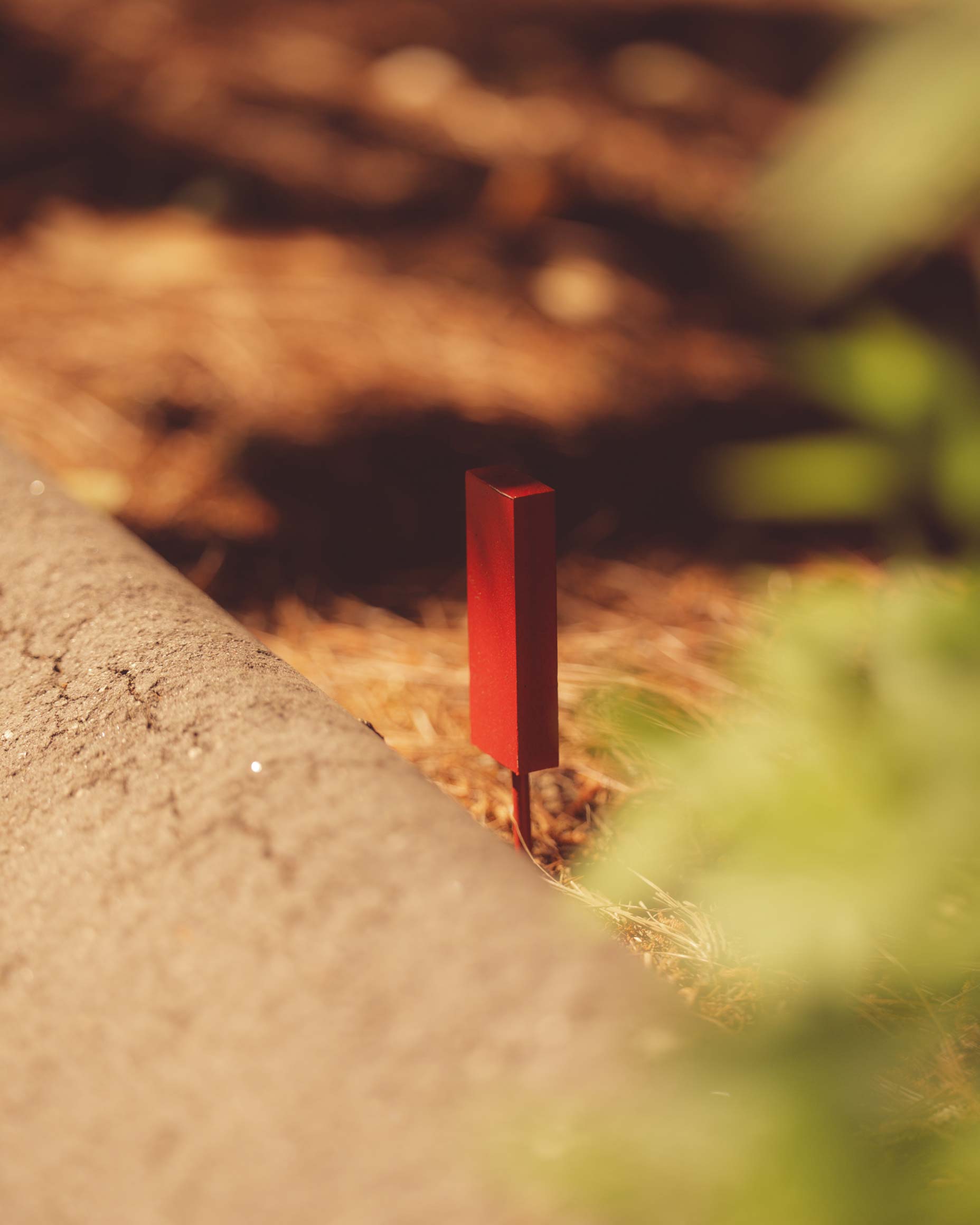 A small, upright red rectangular marker stands in soil beside a concrete edge—reminiscent of the precise markers found at Augusta National golf course—with blurred brown mulch and green foliage in the background.