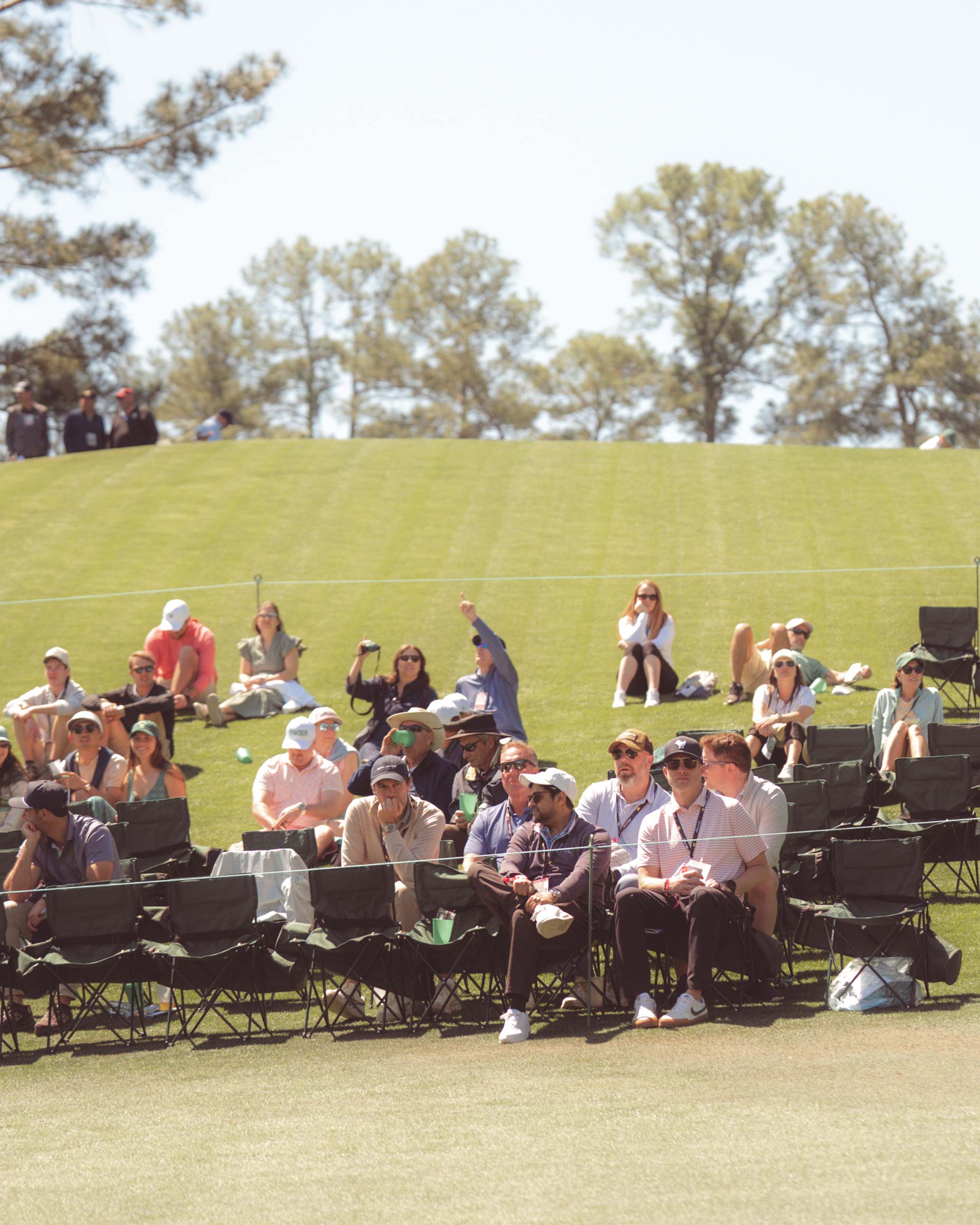 Spectators sit on foldable chairs and grass, watching a golf event on a sunny day. Trees and blue sky are visible in the background, evoking Augusta National vibes. Some wear hats and sunglasses, many relaxed—perhaps after having walked all 18 holes themselves.
