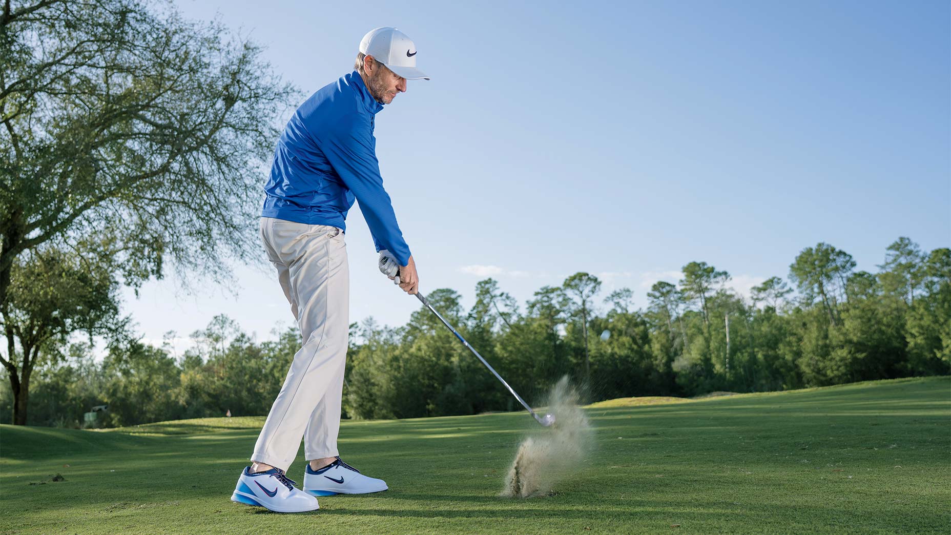A golfer in a blue jacket and white cap takes a powerful swing on the golf course, sending up a small spray of dirt as he strikes the ball, with trees and a clear sky in the background.