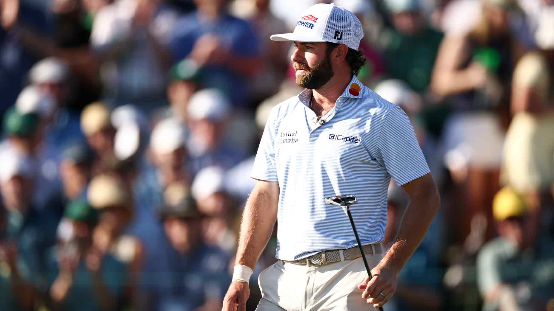 Cameron Young, wearing a white cap and striped shirt, holds a putter on the golf course during the Masters, with a blurred crowd in the background.