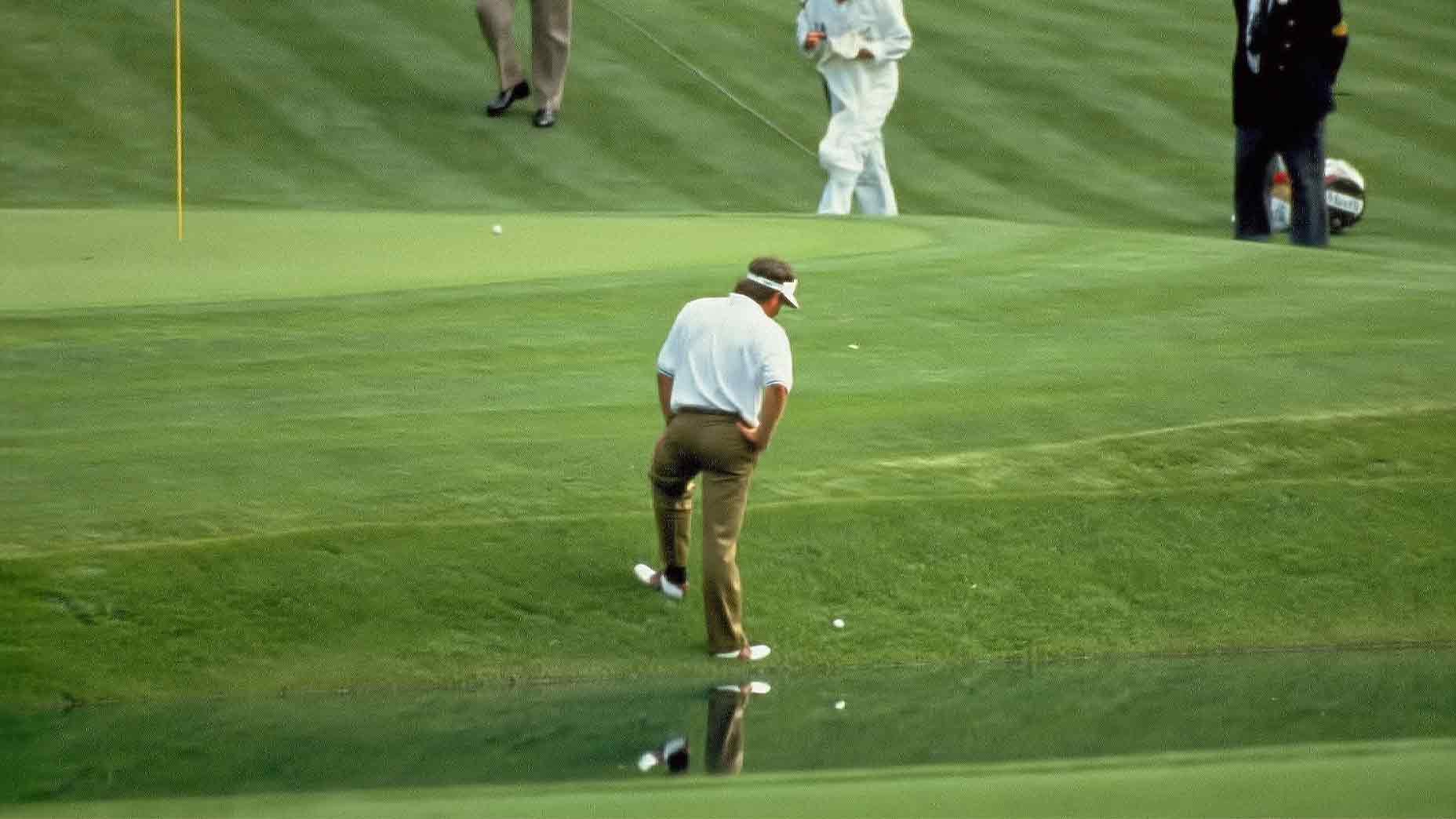 A golfer in brown pants and a white shirt stands near the edge of a water hazard, much like Fred Couples during the 1992 Masters, eyeing his golf ball as if hoping for a bit of divine intervention. Others watch from the manicured grass in the background.