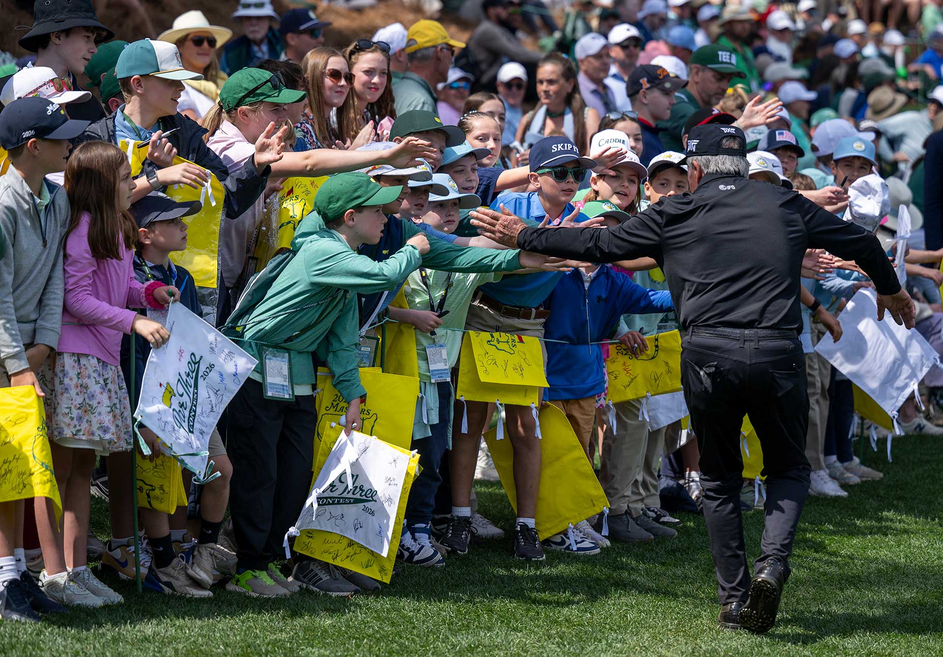 A golfer in black high-fives a crowd of enthusiastic children, many wearing hats and holding yellow bags, as people watch in the background at a Masters par 3 outdoor event.