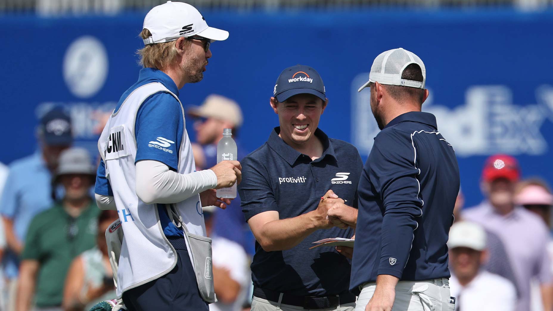 Three men wearing golf attire stand together on a golf course during the 2026 Zurich Classic Saturday tee times; one holds a water bottle, while another smiles and shakes hands with the third man. Spectators and a blue sponsor wall are visible in the background.