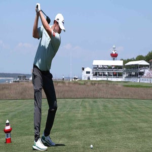 A golfer prepares to swing on a tee box with a golf ball in front of him, as spectators&mdash;possibly awaiting the 2026 RBC Heritage Saturday tee times&mdash;watch from a white pavilion under a clear blue sky.