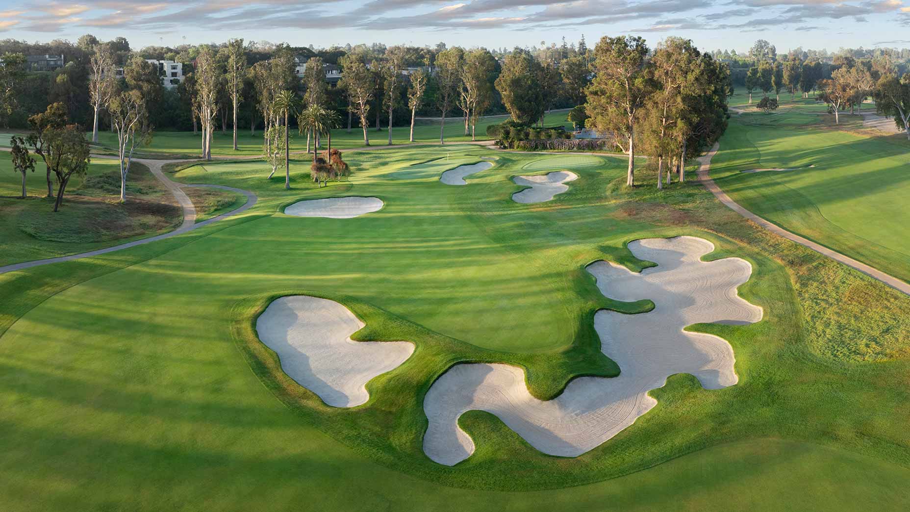 Aerial view of a golf course reminiscent of Riviera, featuring lush green fairways, large sand bunkers, winding paths, and tall trees under a partly cloudy sky—perfect for hosting the U.S. Women's Open.