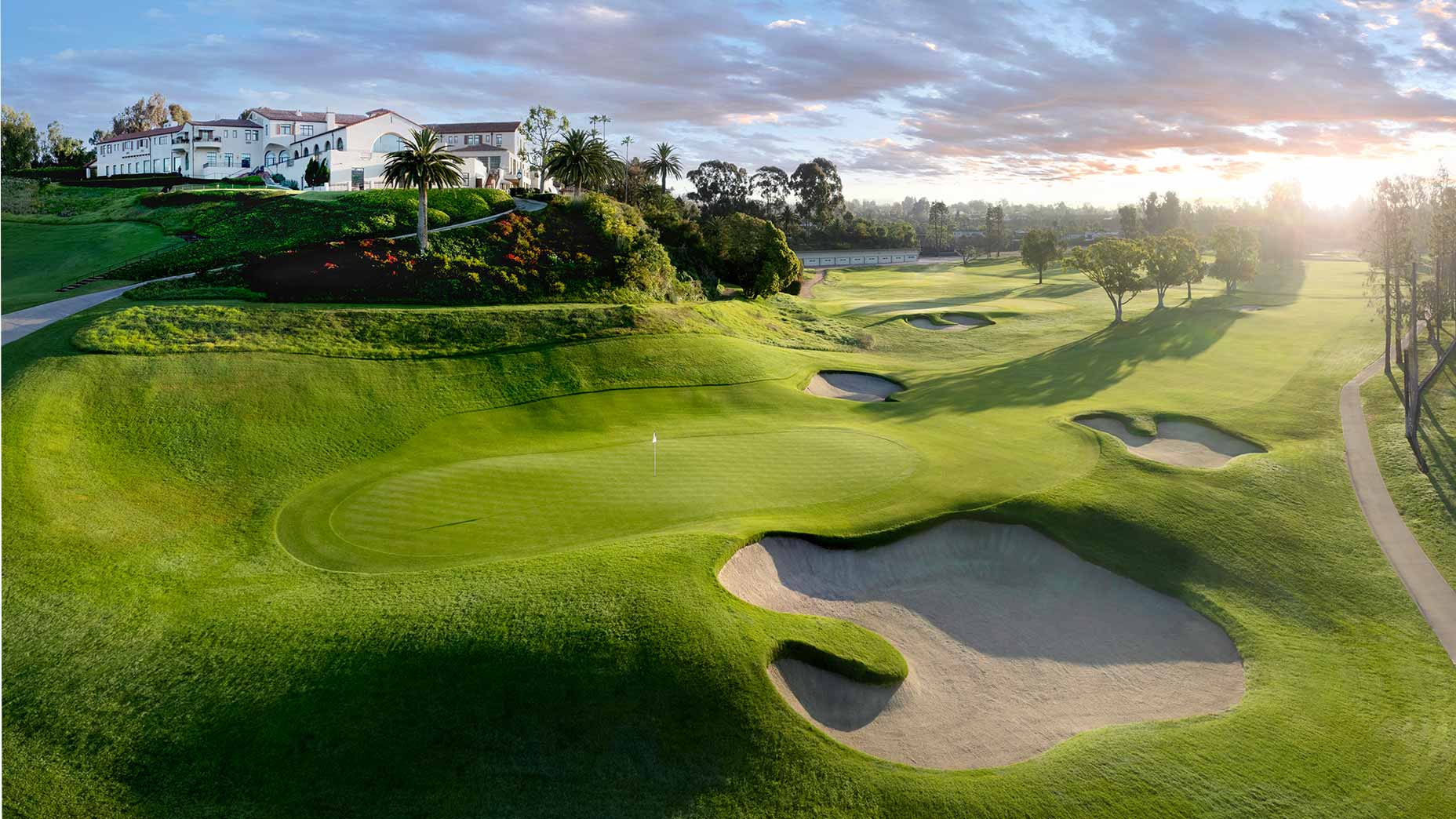 A scenic golf course at sunrise, with smooth green fairways and sand bunkers, evokes the prestige of the U.S. Women's Open—its large white clubhouse on a hill hinting at Riviera’s deeper meaning for golf enthusiasts.