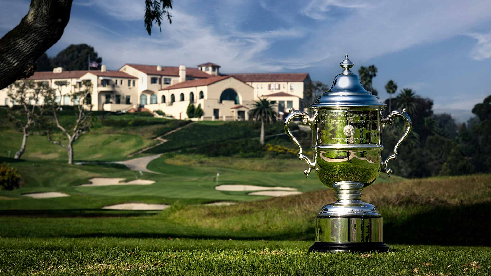 A large silver golf trophy stands on the green grass at Riviera, with a historic clubhouse and the U.S. Women's Open course in the background, all beneath a partly cloudy sky, evoking a deeper meaning of tradition and triumph.