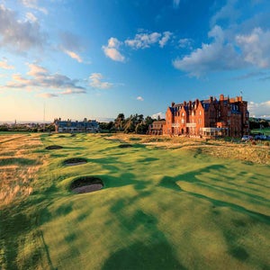 A scenic golf course popular for international golf travel, featuring well-manicured greens and bunkers, bordered by a large historic red-brick building under a blue sky with scattered clouds at sunset. Ideal for your 2027 golf adventures.