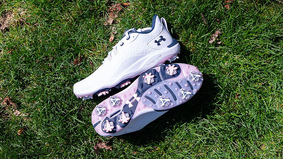A pair of white Under Armour Drive Pro Clone LE golf shoes, one upright and one showing the sole with multiple cleats, rests on green grass in bright sunlight.