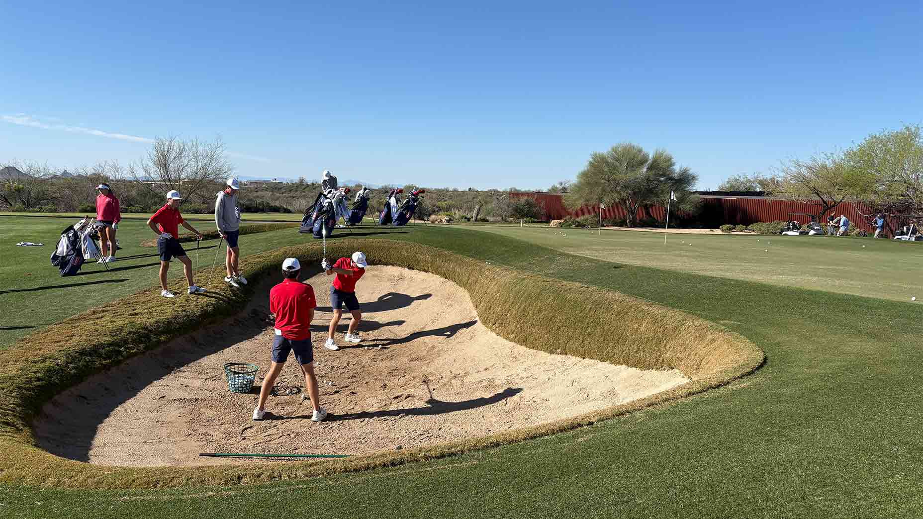 Several young golfers practice hitting balls out of a sand bunker on a sunny, clear day, demonstrating how to improve their technique. Golf bags are lined up in the background as other U.S. National Junior Team members watch nearby.