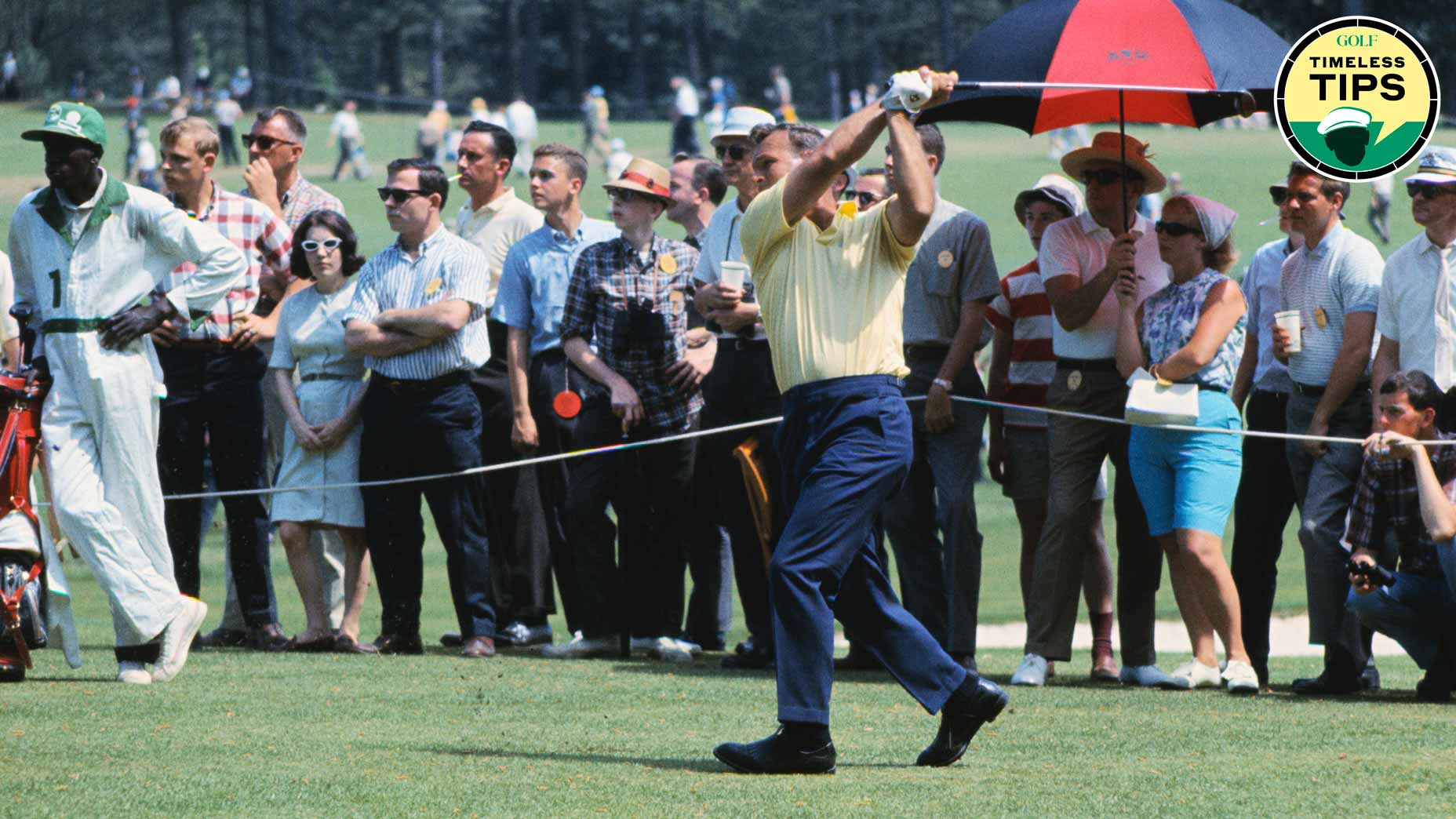 A golfer in a yellow shirt swings his club as a crowd of spectators watches. Some people stand behind a rope, with one holding a red and black umbrella. The scene appears to be at a professional golf tournament.