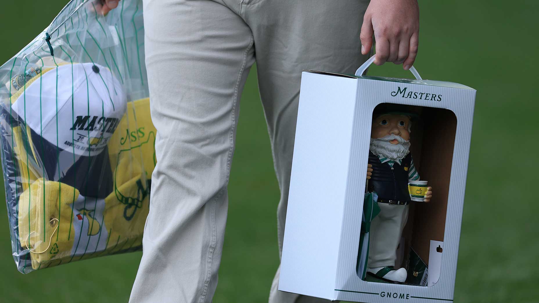 A person holds a Masters gnome in a box with one hand and a clear bag filled with Masters merchandise, including hats and shirts, in the other hand.