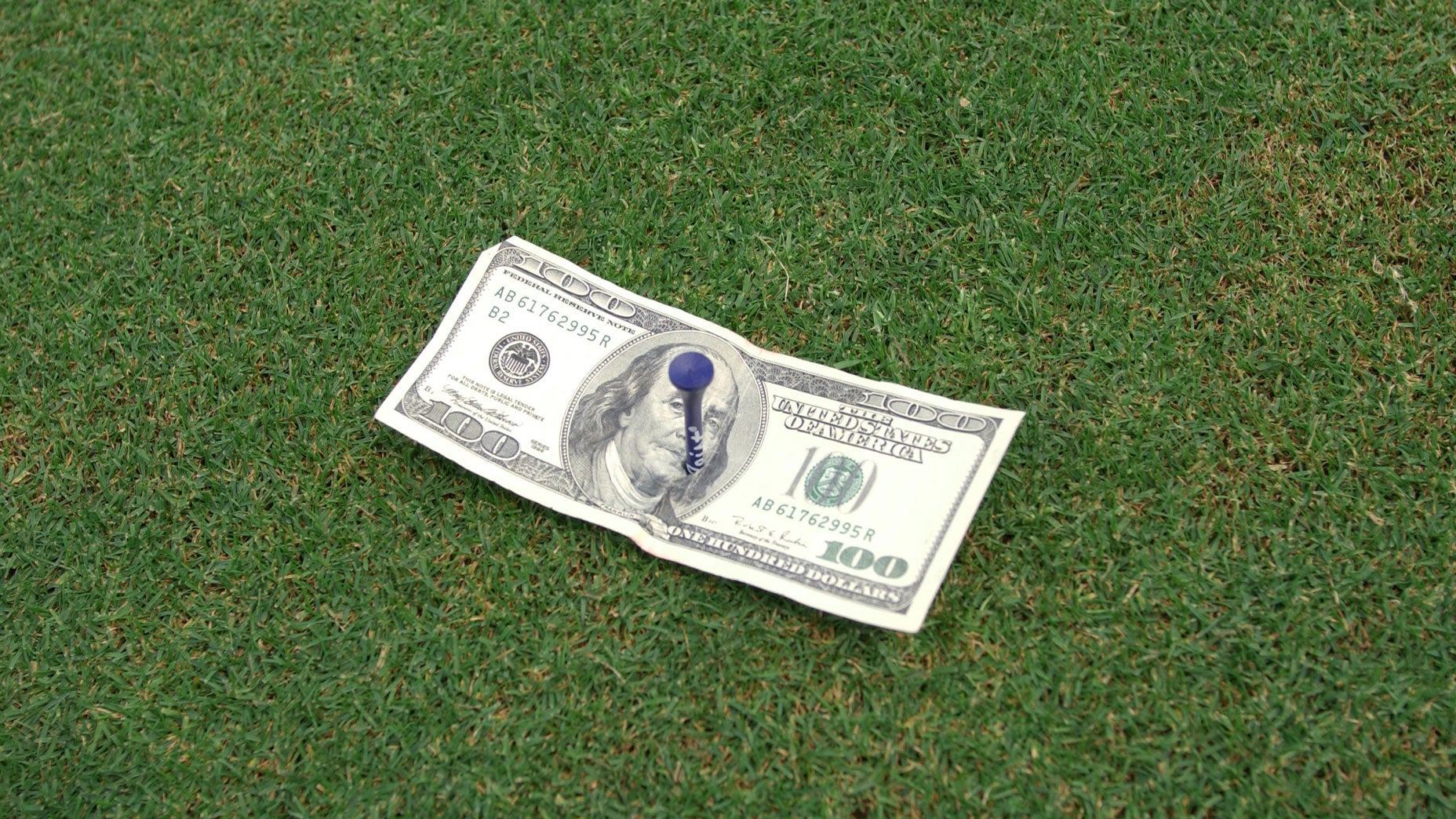 A $100 bill with a blue golf tee placed on top of it lies on green grass, resembling a putting green—a clever nod to the cost of green fees.