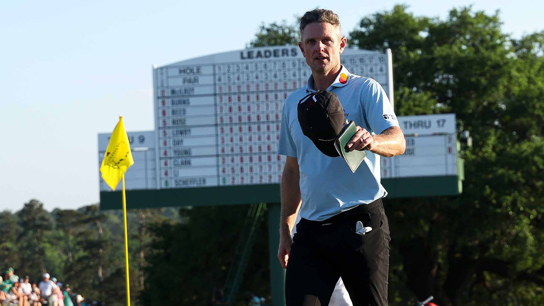 Justin Rose holds his cap and scorecard at the Masters near a yellow flag, with a large leaderboard and trees visible in the background.