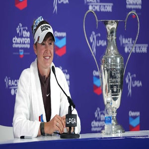 A female golfer, Nelly Korda, in a white jacket and cap sits at an LPGA press conference table, smiling beside a large Chevron Championship trophy after her Chevron win, with a water bottle in front of her and a blue backdrop behind.