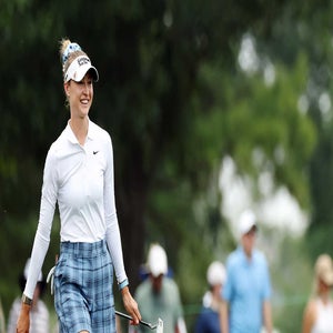 Nelly Korda in a white long-sleeve shirt and blue plaid pants smiles while holding a putter on the golf course during the 2026 Chevron Championship, with blurred trees and people in the background.