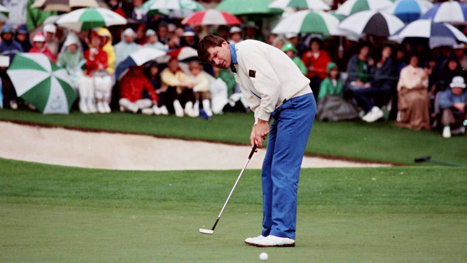 A golfer in blue pants and a white sweater putts on the green at Augusta National, as a crowd of spectators with umbrellas watches from near the sand bunker—capturing one of the scariest shots faced by any Masters winner.