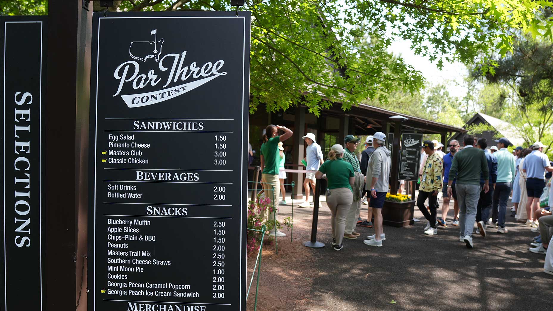 A menu board listing sandwiches, beverages, snacks, and Masters merchandise stands outdoors near trees, with people waiting in line by a wooden building in the background.