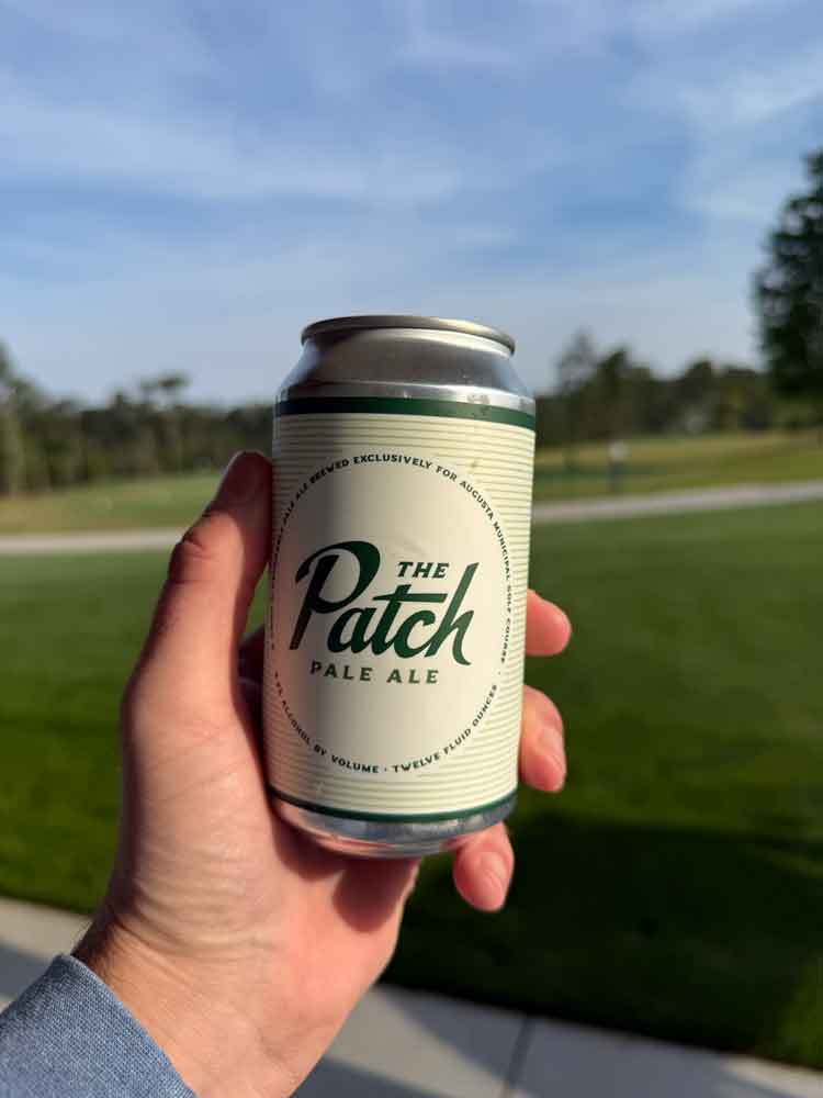 A hand holding a can of The Patch Pale Ale beer outdoors, with a grassy lawn reminiscent of Augusta National and trees in the background under a clear sky.