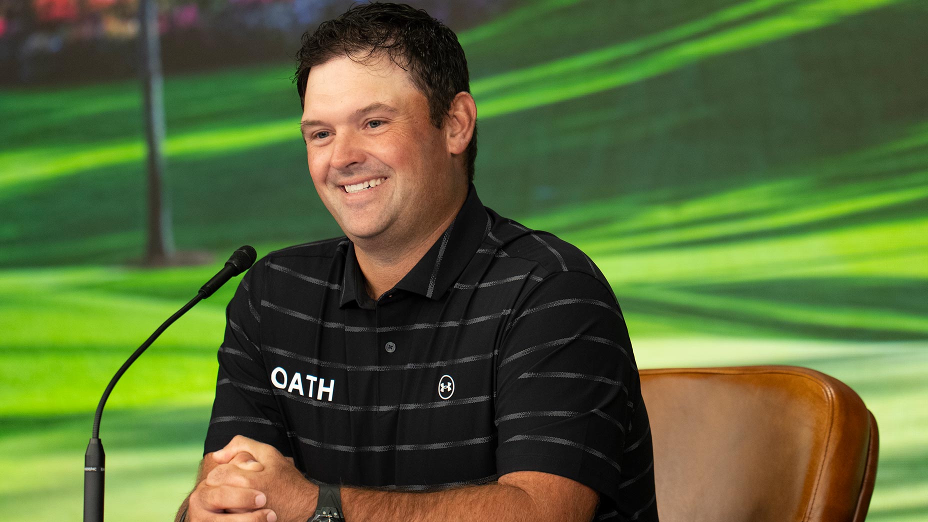 A man wearing a black striped polo shirt with the word OATH on it sits smiling at a table in front of a microphone, with a green, outdoor background behind him—reminiscent of Patrick Reed at the Masters.