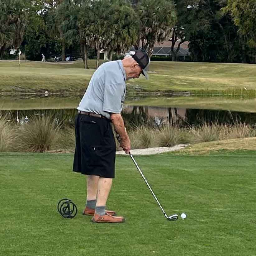 An elderly man in a cap and shorts prepares to hit a golf ball on a green course in Boca Raton, with a pond and trees in the background, enjoying his golf trip.