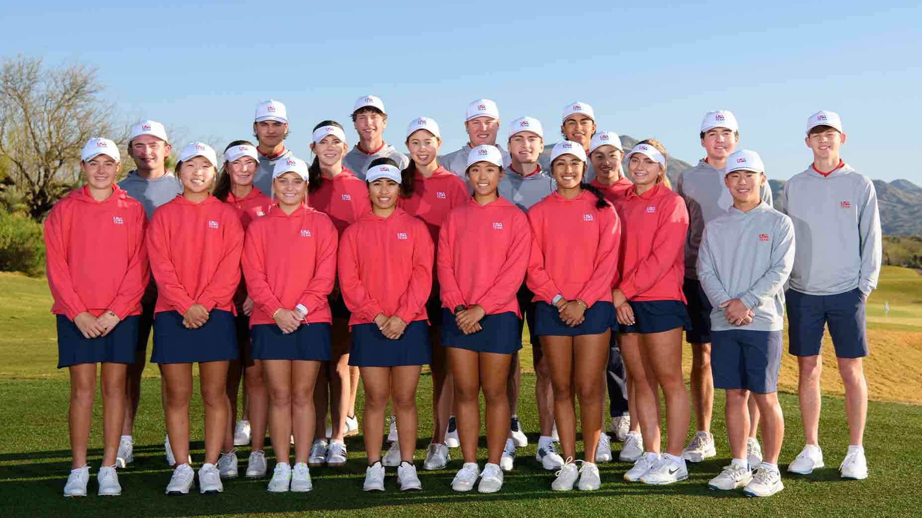 A group of young golfers from the U.S. National Junior Team, wearing matching red, gray, and white uniforms, stand together outdoors on a golf course with clear skies and mountains in the background, smiling after practice for a team photo.
