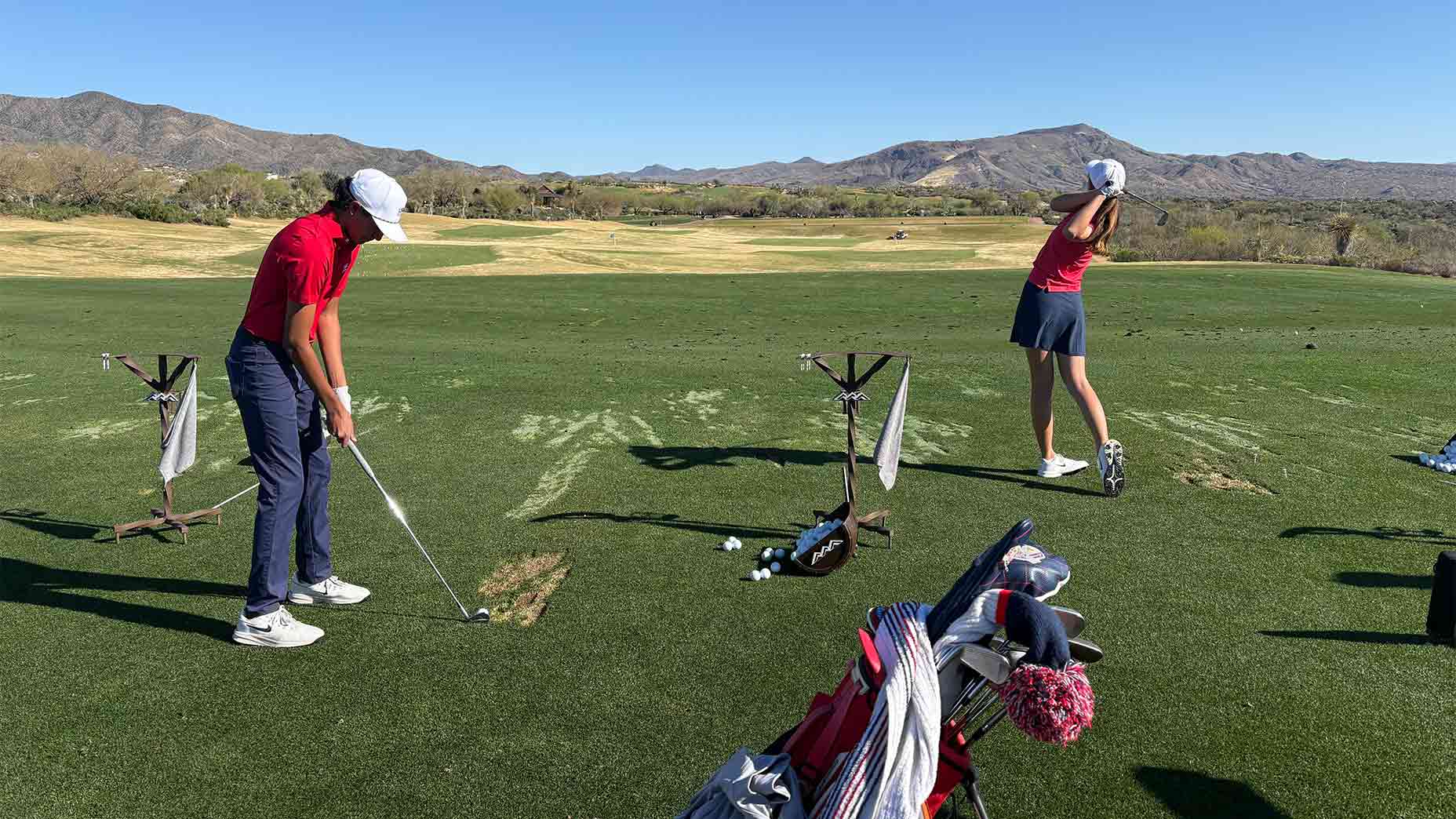 Two golfers practice their swing on a driving range with mountains behind them. Both wear red shirts and white hats—one mid-swing, the other preparing to hit. Their focus mimics a U.S. National Junior Team training session among golf bags and scattered balls.