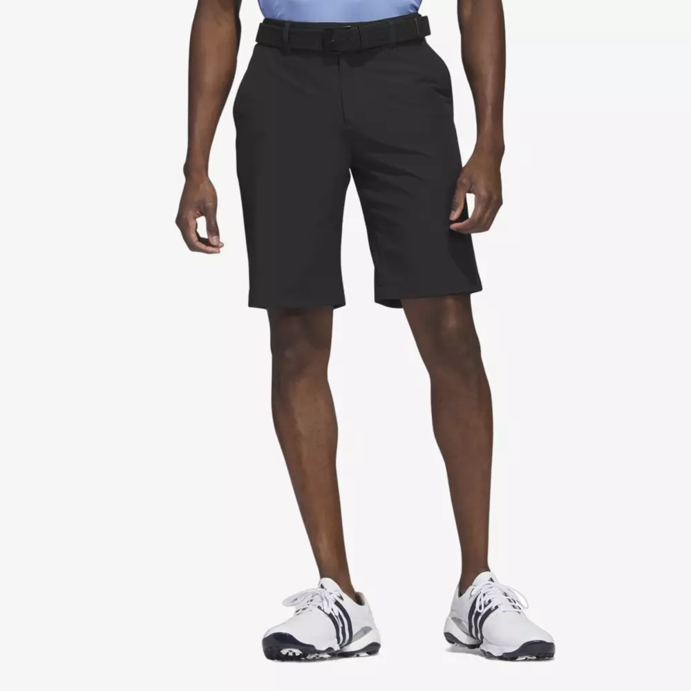 A person wearing black knee-length shorts, a light blue shirt, a black belt, and white athletic shoes with black accents stands against a plain white background, ready to pack one of the best golf travel bags for their next adventure.