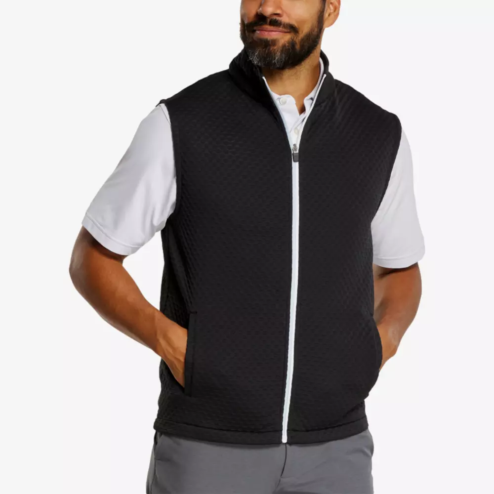 A man with a beard wearing a white polo shirt, gray pants, and a black zip-up vest showcases stylish comfort perfect for the green—ideal for those seeking the best golf deals for $100 or less.