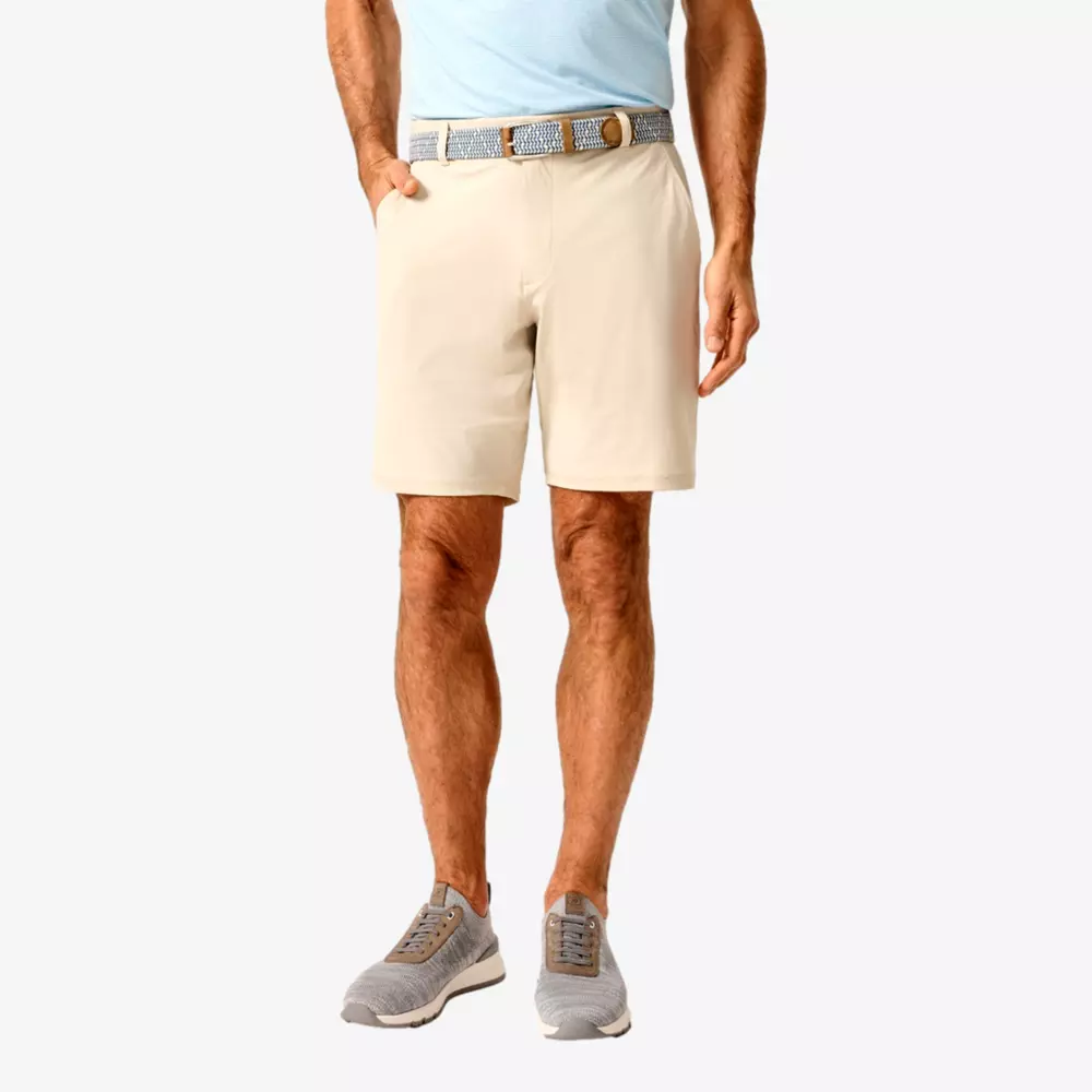 A man wearing beige shorts, a blue belt, a light blue shirt, and gray sneakers stands with one hand in his pocket against a plain light background—dressed perfectly for browsing the best golf travel bags.