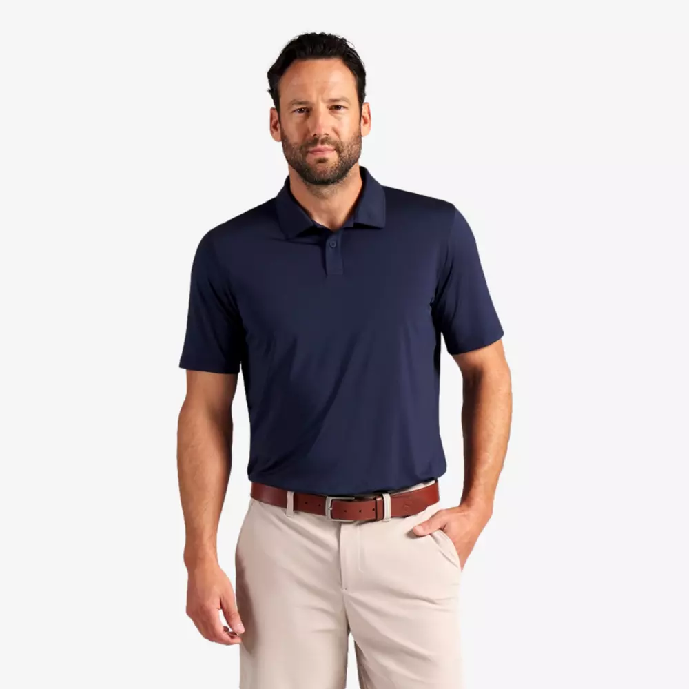 A man with a beard wearing one of the best golf polos&mdash;a navy blue short-sleeve polo shirt&mdash;paired with beige pants and a brown belt, stands with one hand in his pocket against a plain light background.