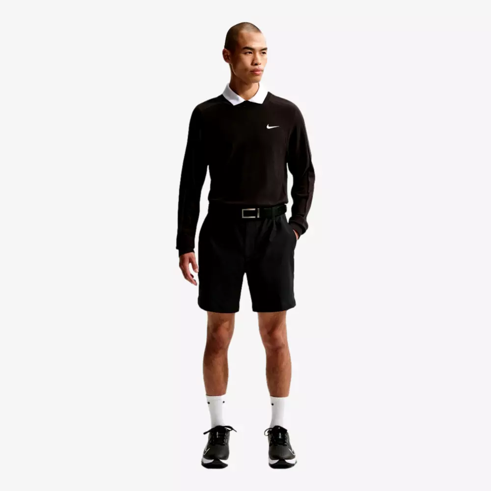 A man stands facing forward, wearing a black long-sleeve Nike shirt with a white collar, black shorts, a black belt, white socks, and black athletic shoes—ready for the course or to pack up with the best golf travel bags against a plain white background.