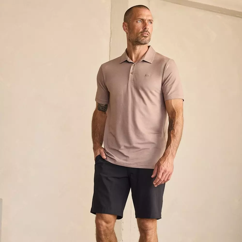 A man stands indoors wearing a light brown polo shirt and black shorts, perhaps planning his next trip and considering the best golf travel bags. He looks to his left with a neutral expression, his right hand relaxed at his side and his left hand lightly touching his thigh.