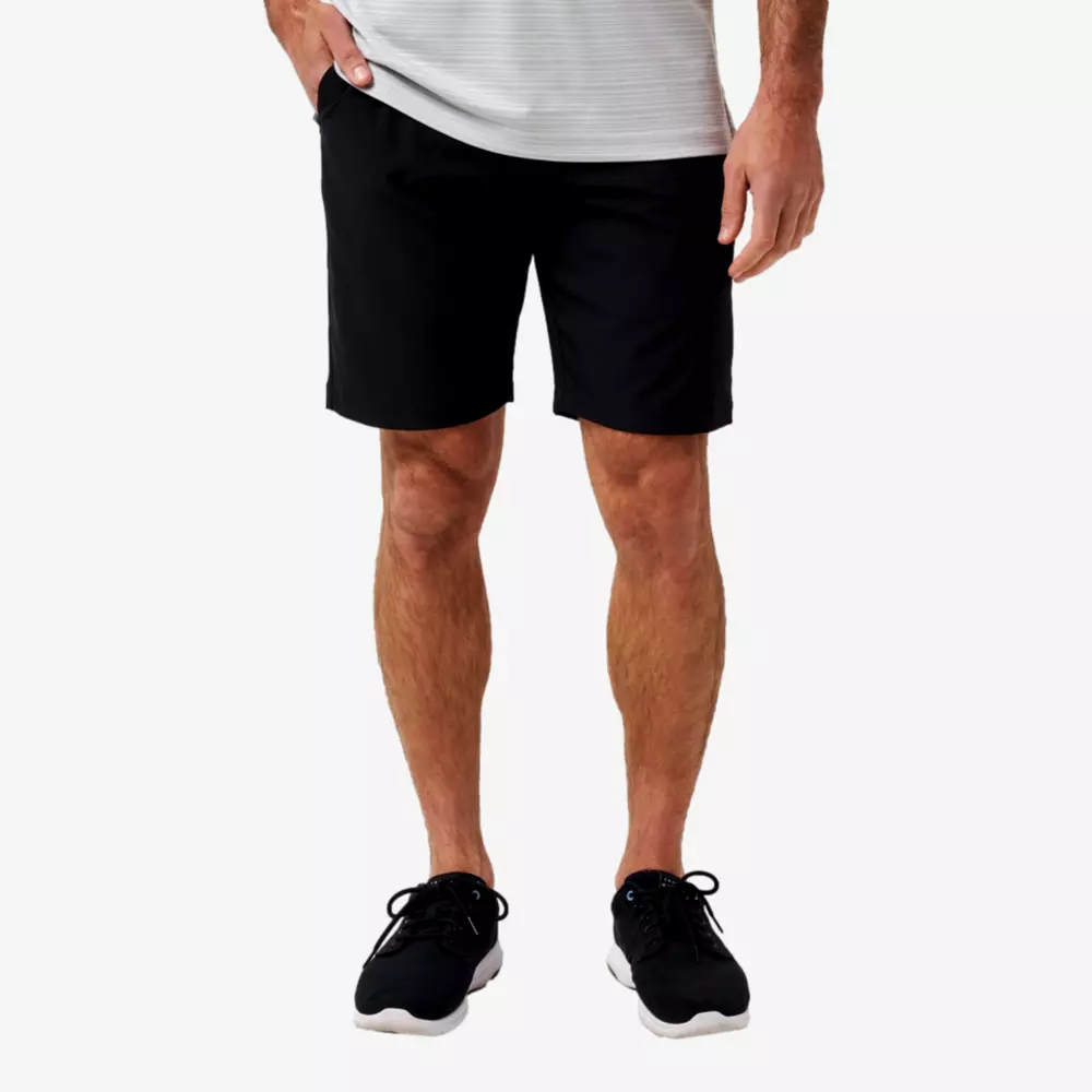A man wearing a light gray striped shirt, black shorts, and black athletic shoes stands with one hand in his pocket against a plain white background, looking ready to choose from the best golf travel bags for his next trip.