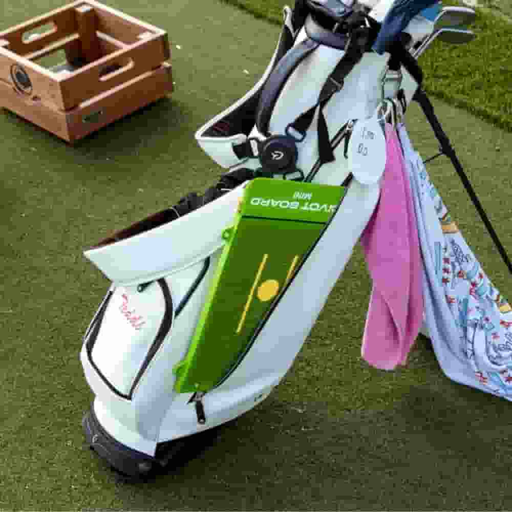 A contender for the best golf bags 2026, this white golf bag with clubs, a green alignment board, pink towel, and patterned cloth stands on grass beside a wooden crate.