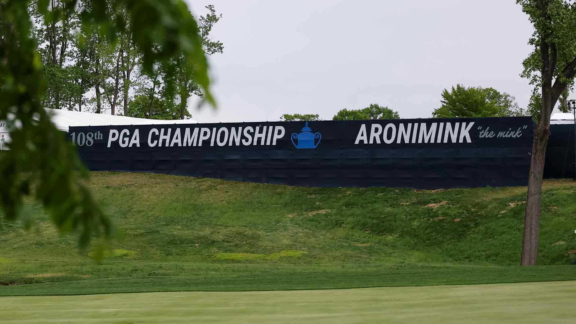 Large 2026 PGA Champioinship sign behind a golf course with a blue trophy icon, surrounded by grass and partially framed by tree leaves.
