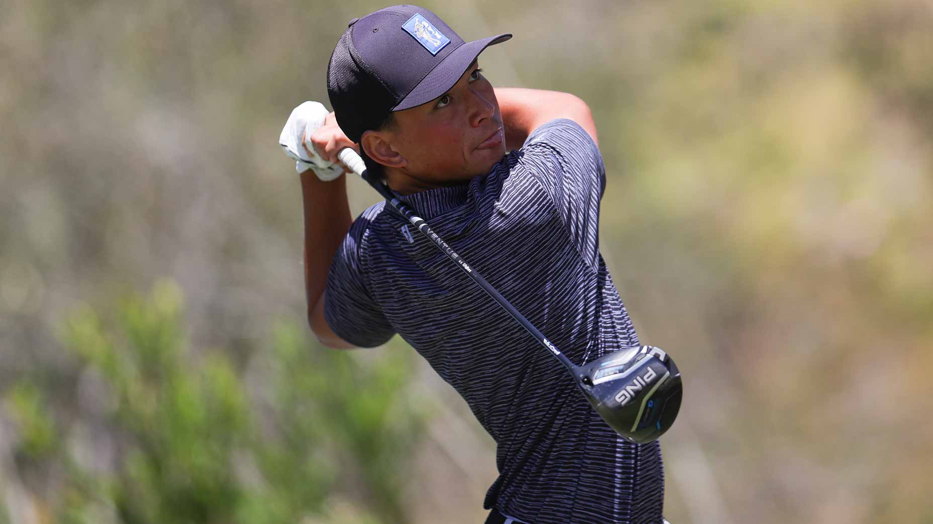 A golfer wearing a striped shirt and black cap follows through on a golf swing outdoors, holding a Ping driver, with greenery blurred in the background at the BYU course.