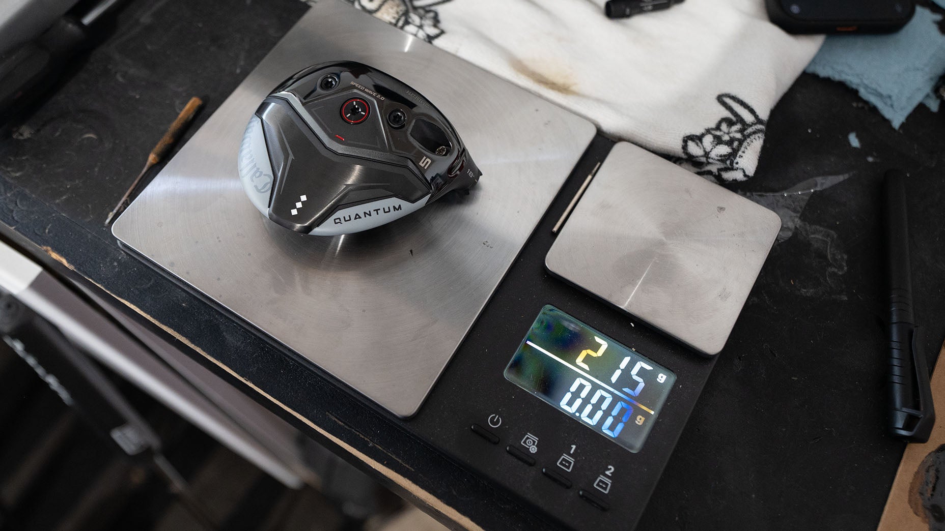 A metallic golf club head labeled Quantum sits on a digital scale, displaying a weight of 215 grams. A golf shaft tip and manufacturer tools are visible in the background on the dark surface, alongside a white cloth.