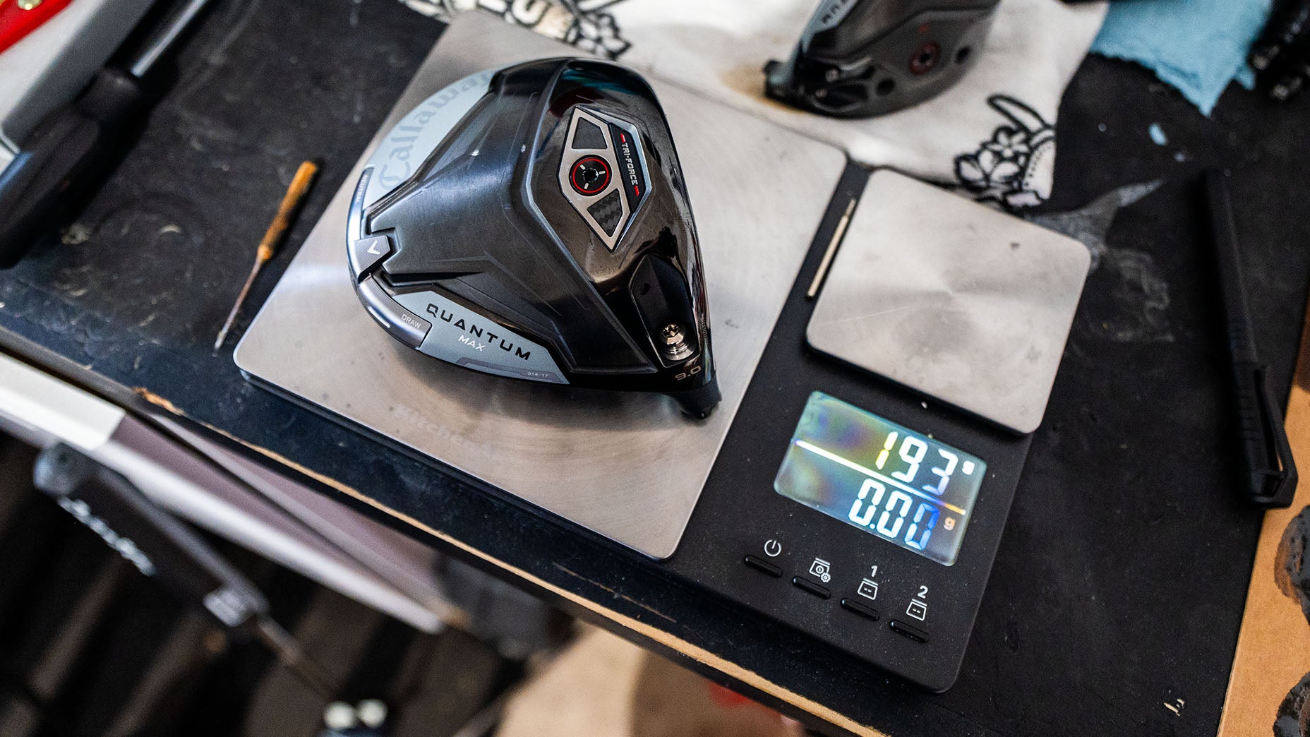 A black golf club head labeled Quantum rests on a digital scale showing 193.7 grams, while nearby golf shafts and tools hint at the precision of a dedicated manufacturer’s workspace.