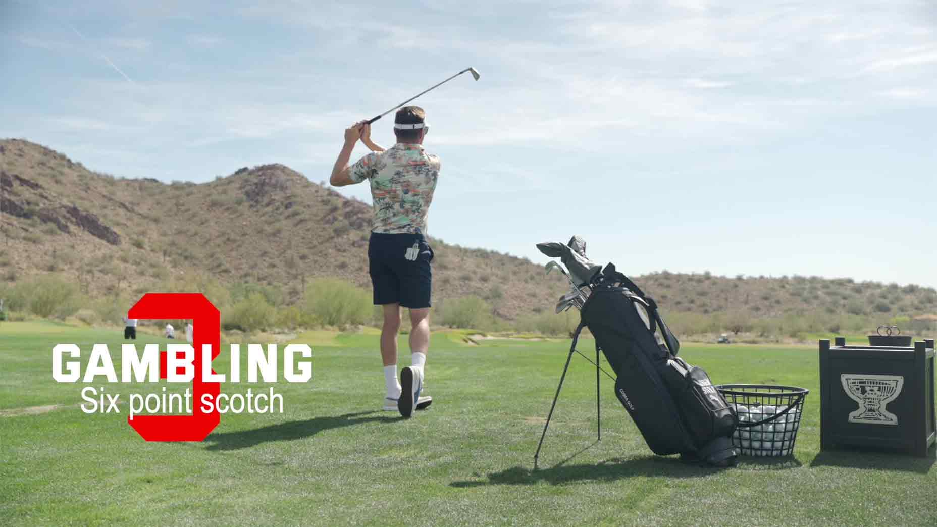 A golfer swings a club on a grassy course with desert hills in the background. Golf bag and basket of balls nearby. Text reads: GAMBLING Six point scotch, inspired by Drew Stoltz, with a large red G behind the word GAMBLING-game advantage.