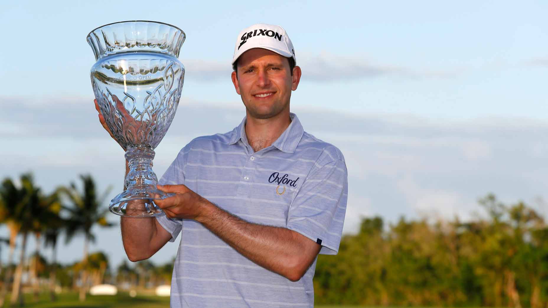 A retiring pro in a striped golf shirt and cap smiles while holding a large crystal trophy outdoors, with palm trees and greenery behind him—celebrating his career and sharing his golf secrets from years on the PGA Tour.