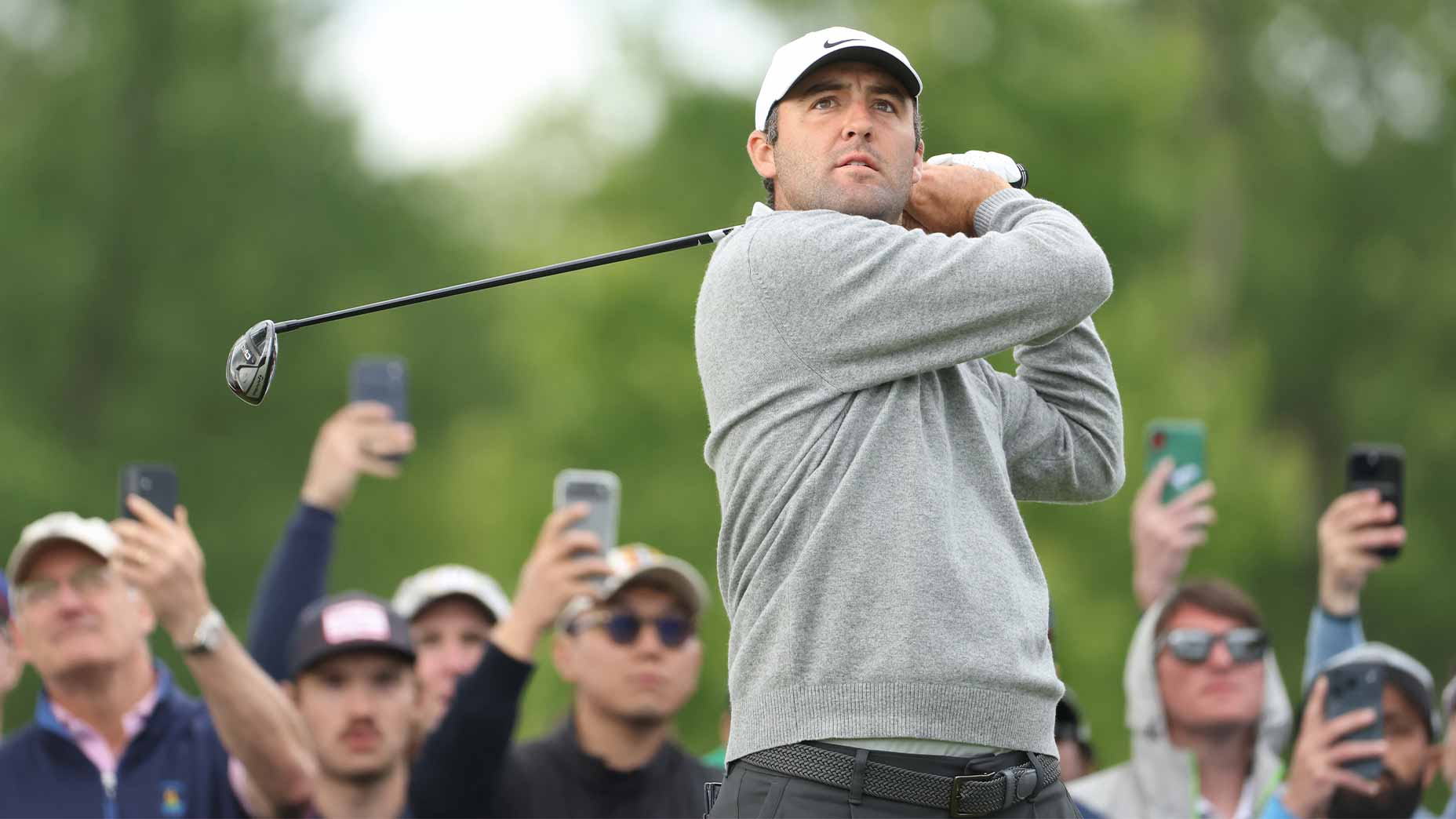 A golfer in a gray sweater and white cap follows through on a swing at the PGA Championship, while a group of spectators behind him watch and take photos with their phones. The background is green and outdoors.