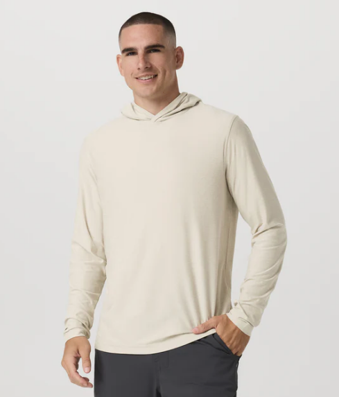 A man with short dark hair, wearing a light beige Vuori long-sleeve hooded shirt and dark pants, stands against a plain white background, smiling with one hand in his pocket, reflecting brand loyalty.