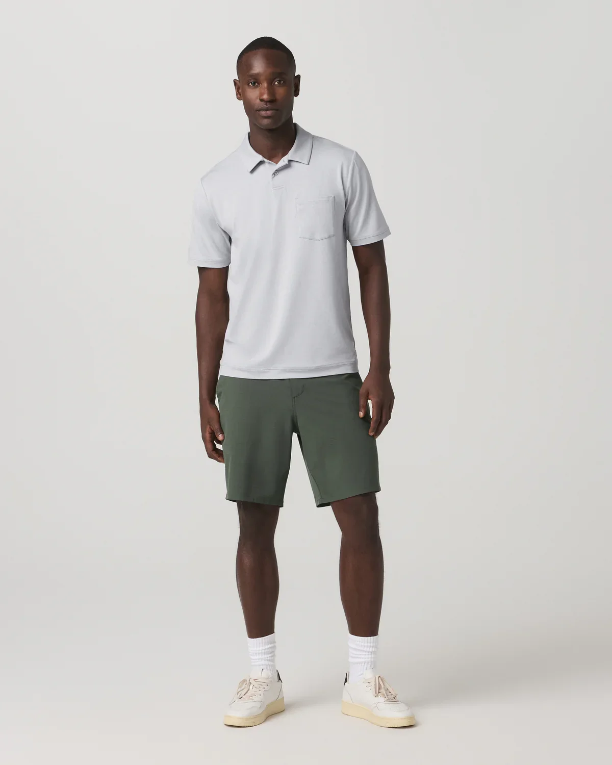 A man stands against a plain light background, wearing a light gray polo shirt, olive green shorts, white socks, and beige sneakers—dressed as if ready to browse the best golf travel bags with his relaxed expression and posture.